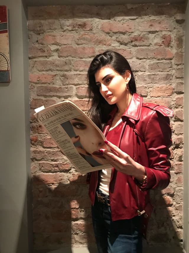 "Güzellik Yayında" başlığı olan bir gazete okuyan kırmızı ceketli kadın