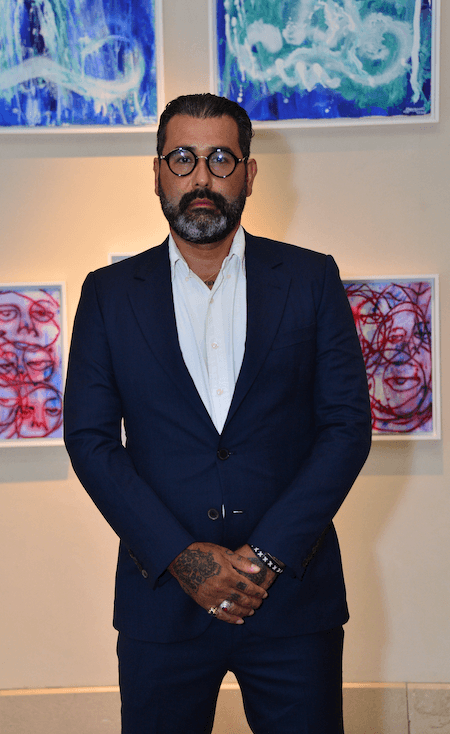 Mavi takım elbiseli, gözlüklü, sakallı adam sanat tablolarının önünde duruyor