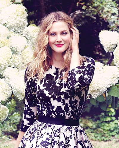 Drew Barrymore siyah beyaz çiçekli elbiseyle beyaz ortancalar arasında gülümsüyor