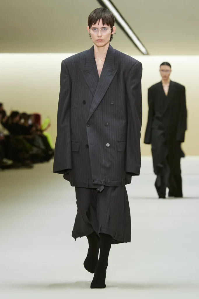 Oversize takım elbiseli model podyumda yürüyor, arkada başka model var