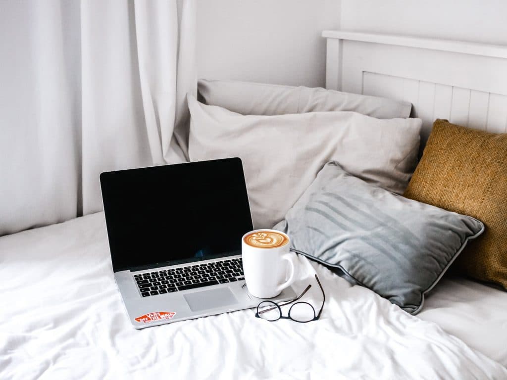 Beyaz yatakta laptop latte art'lı kahve ve okuma gözlükleri