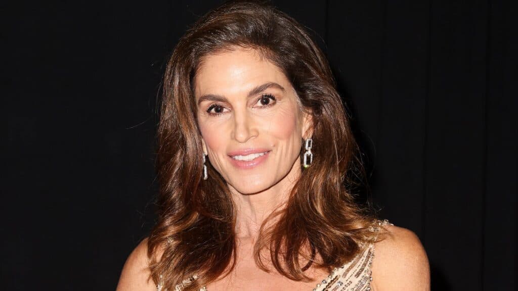 Cindy Crawford kahverengi saçlı, küpeli, koyu arka plan önünde gülümsüyor
