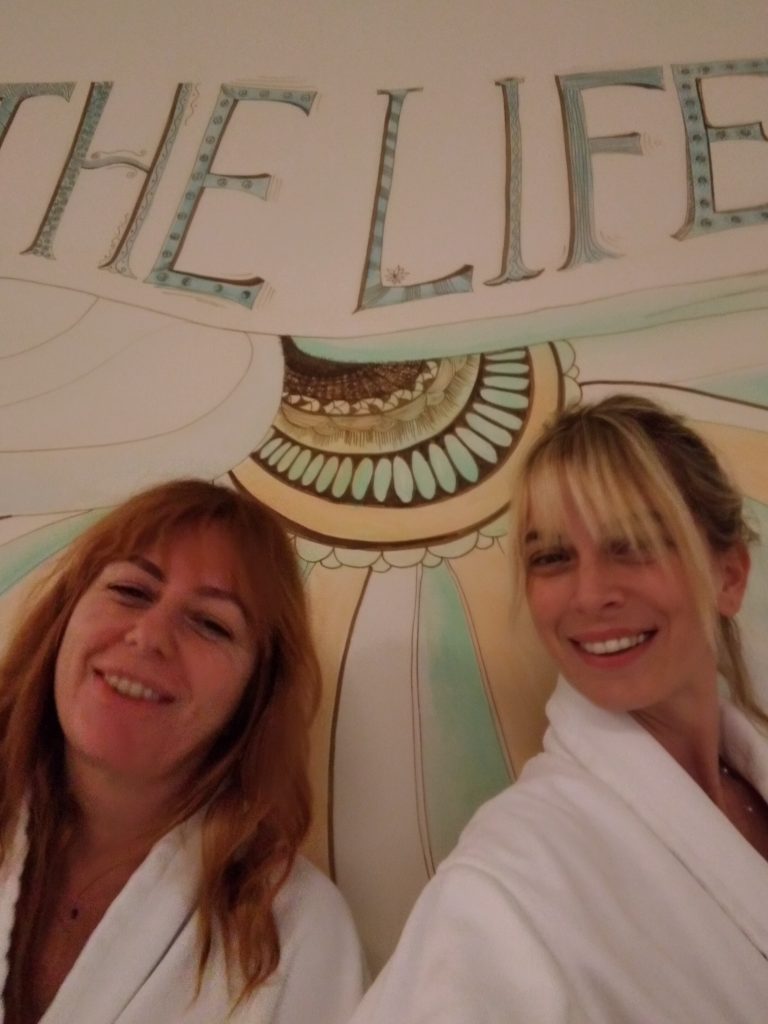 Bornozlu iki kadın "THE LIFE" yazılı duvarda neşeyle gülümsüyor