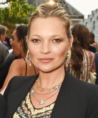 Kate Moss, sarı saçları toplanmış, zarif kolye ve küpeleriyle siyah ceketli