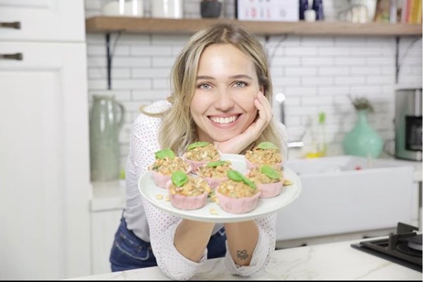 Mutfakta gülümseyen kadın, elinde fesleğen yapraklı muffin tabağı tutuyor