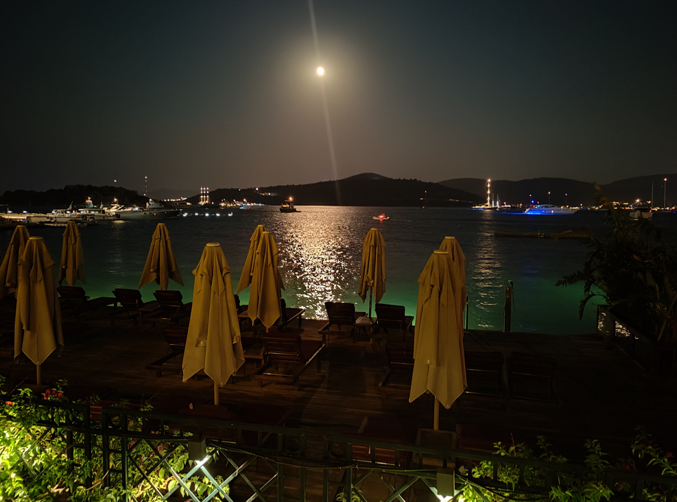 Gece ay ışığı vuran deniz kenarı şemsiyeler tekneler uzakta dağlar