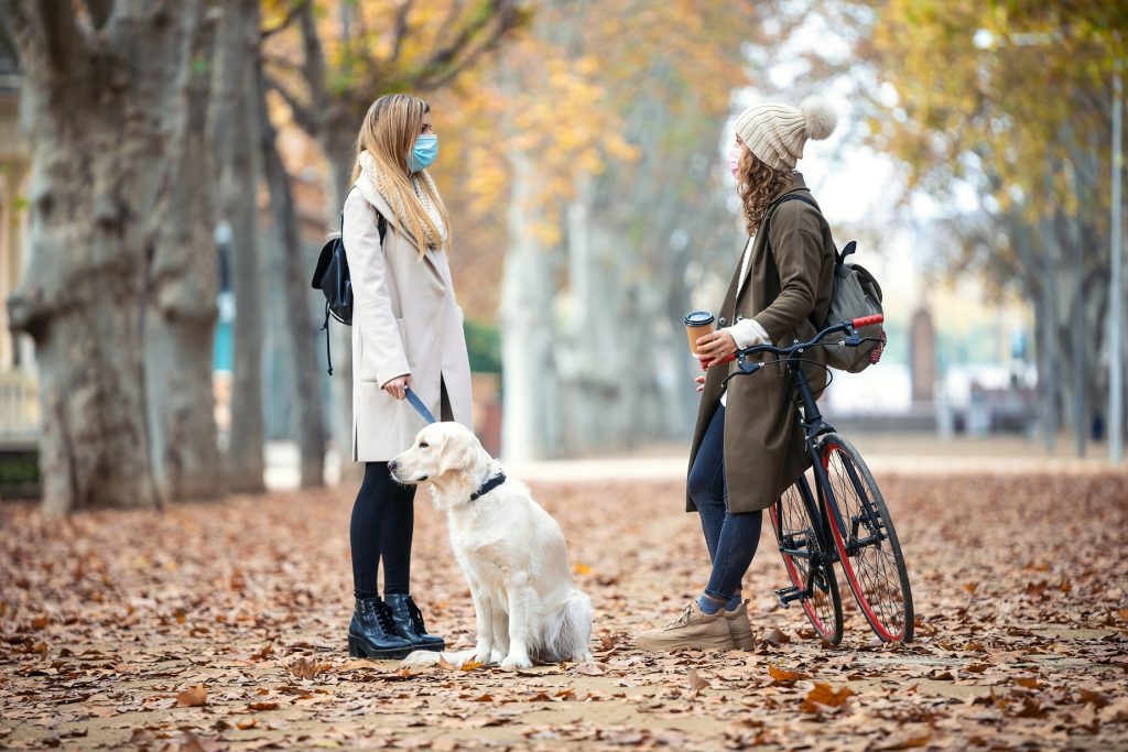 Sonbaharda yapraklı parkta maskeli kadın köpeğiyle bisikletli arkadaşıyla sohbet ediyor