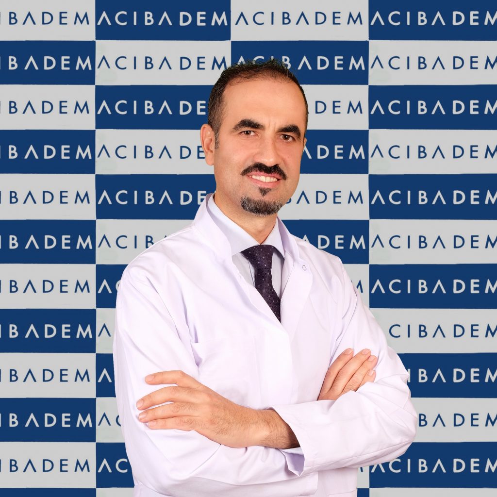 Beyaz önlüklü gülümseyen doktor, Acıbadem logosu önünde duruyor