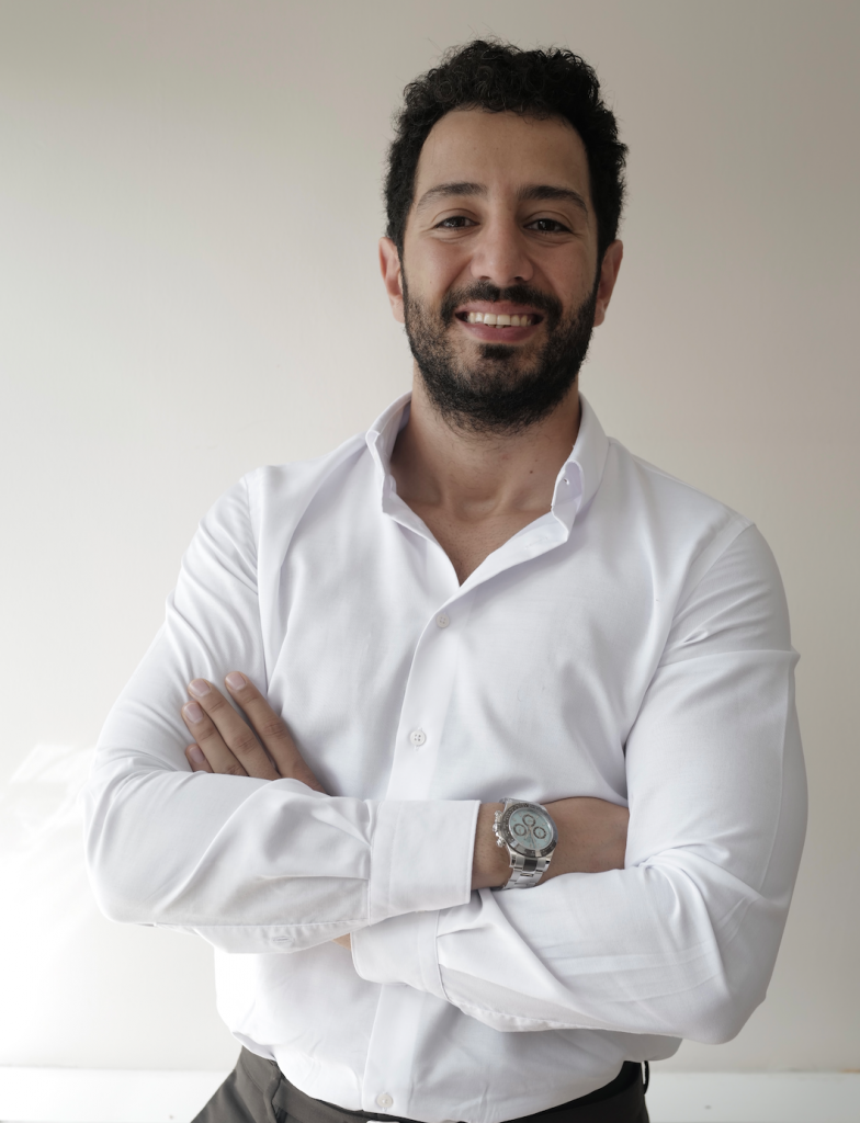 Beyaz gömlekli sakallı adam kollarını kavuşturmuş neşeyle gülümsüyor
