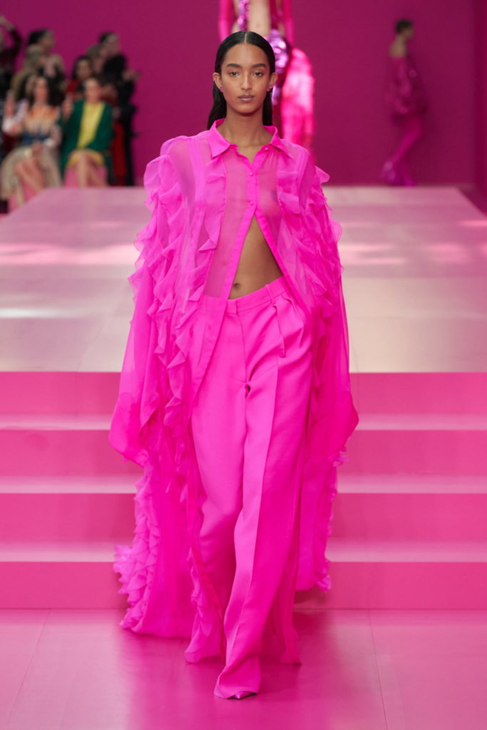 Parlak pembe fırfırlı şeffaf gömlek ve pantolon giyen model podyumda yürüyor