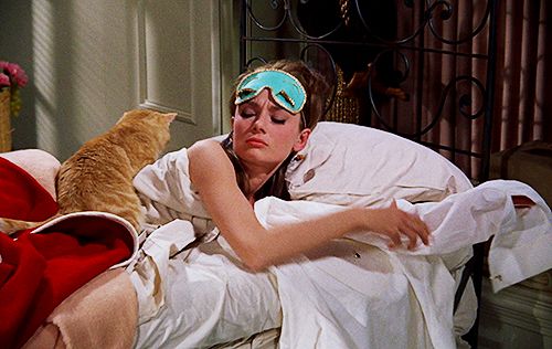 Uykulu Audrey Hepburn yatakta uyku maskesi ve turuncu kedisiyle