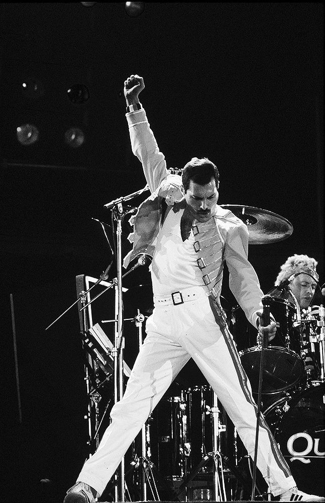 Freddie Mercury sahnede beyaz kıyafetiyle kolunu havaya kaldırmış