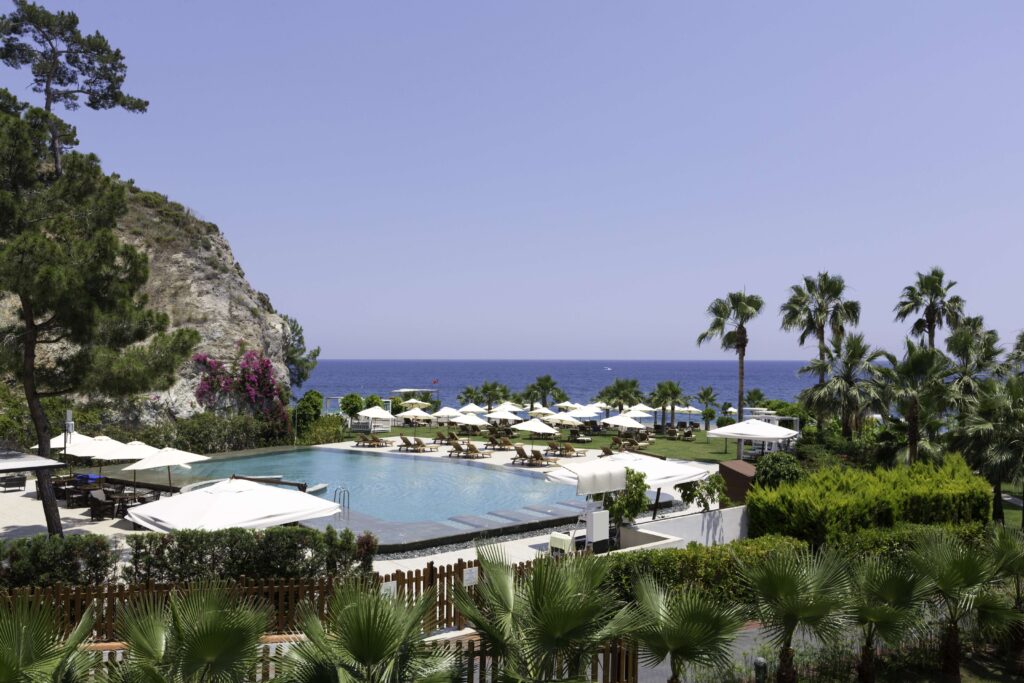 Kayalık yamaç, masmavi deniz, havuz, palmiye ağaçları ve şezlonglu tatil mekanı
