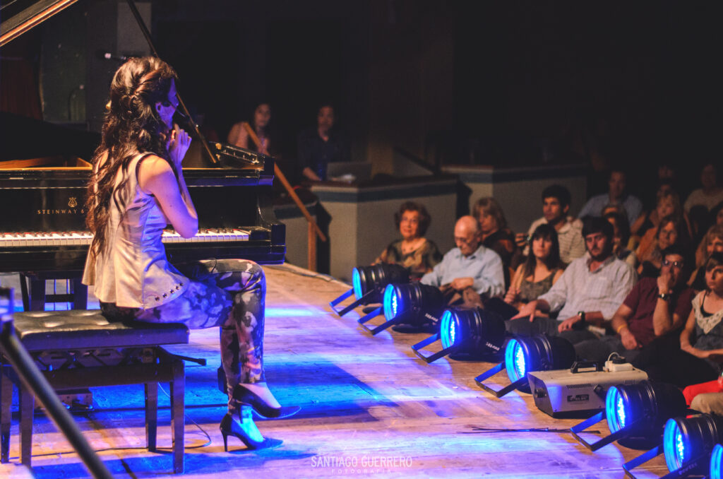 Kadın piyanist sahnede, seyirciler ve mavi spot ışıklarıyla canlı performans