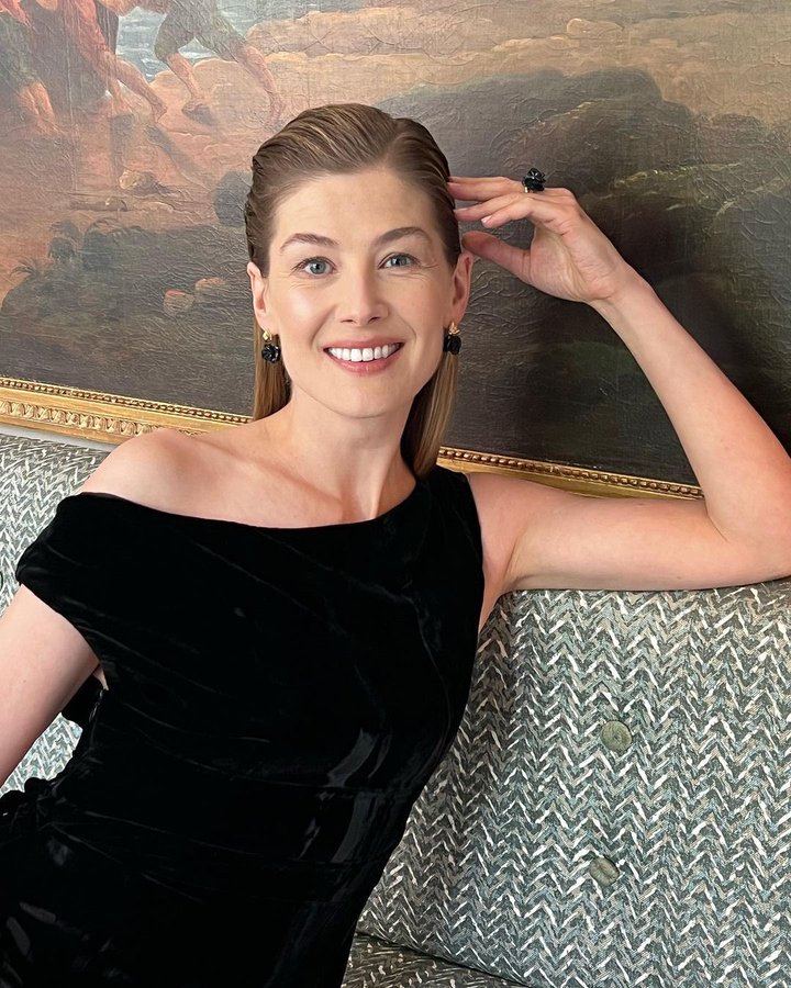 Rosamund Pike siyah kadife elbiseli, tablo önünde kanepeye yaslanmış gülümsüyor