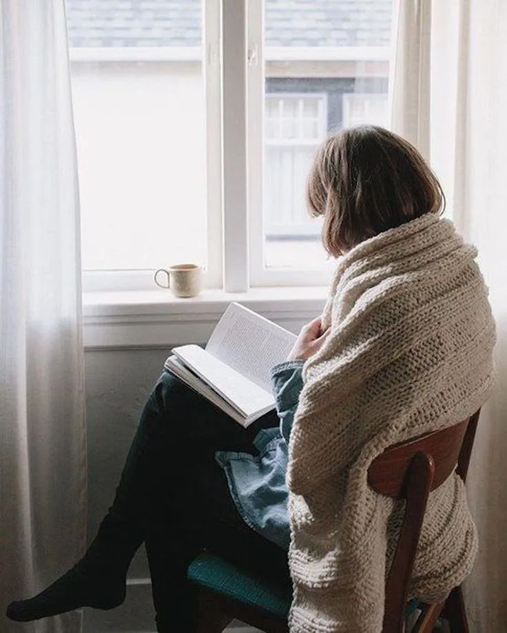 Pencere önünde battaniyeye sarılıp kitap okuyan kişi rahat anlar yaşıyor