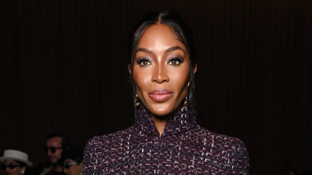 Naomi Campbell koyu renkli, dokulu bir ceketle doğrudan kameraya bakıyor