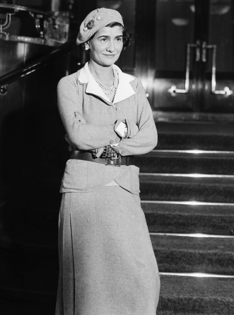 Coco Chanel gri takım elbise ve şapkayla merdivenlerde duruyor