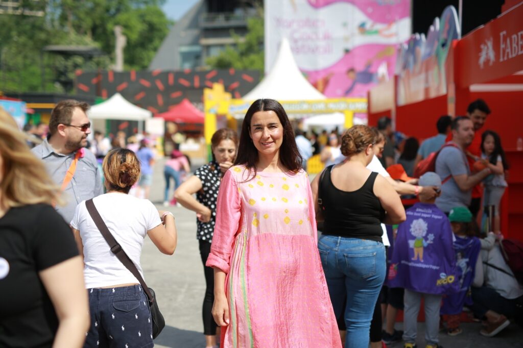 Açık hava etkinliğinde etrafı insanlarla dolu pembe elbiseli kadın gülümsüyor