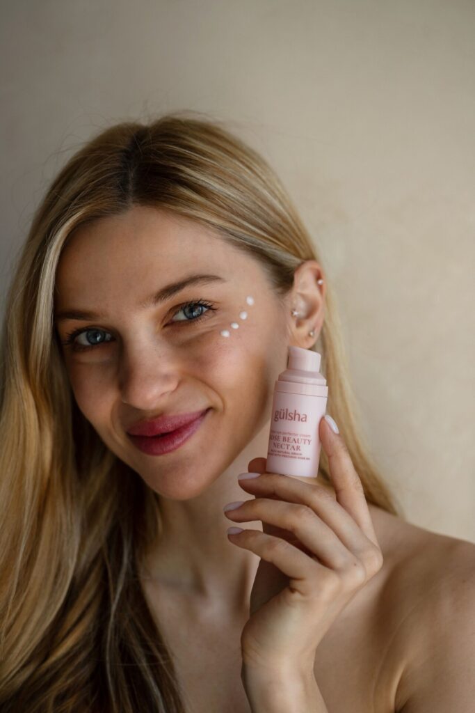 Gülümseyen sarışın kadın yüzünde kremle Gülsha Rose Beauty Nectar şişesini tutuyor