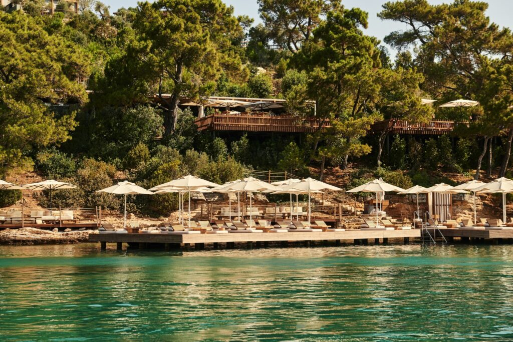 Şemsiyeli iskele, turkuaz deniz ve ormanlık yamaçta tesis
