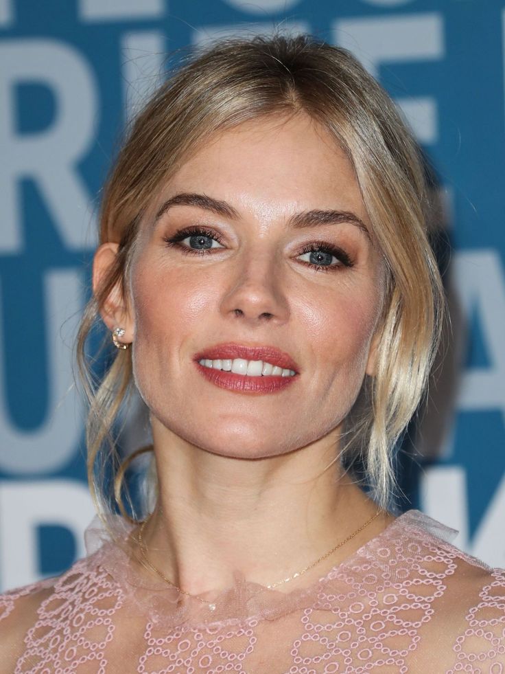 Sienna Miller pembe elbiseyle gülümsüyor, mavi arka plan önünde