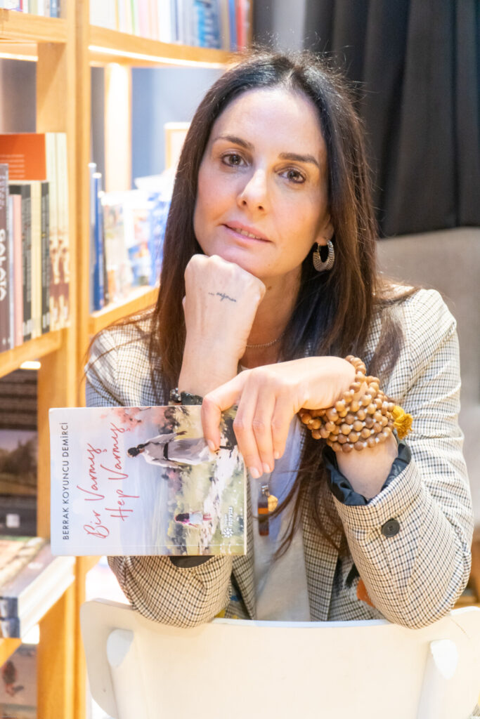 Kitap tutan, kameraya bakan, koyu saçlı bir kadın kitapçı ortamında