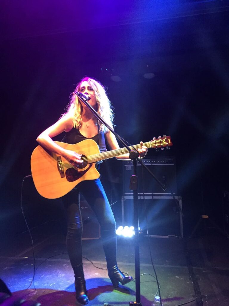 Mor ışıklı sahnede akustik gitar çalıp şarkı söyleyen uzun saçlı kadın