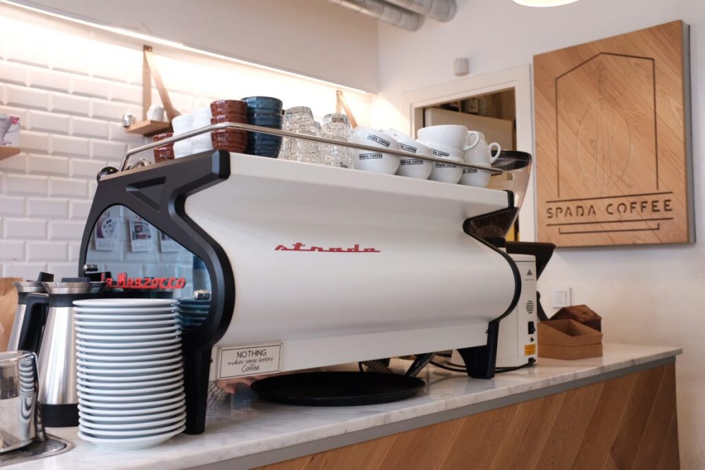 Beyaz La Marzocco Strada kahve makinesi, bardaklar ve Spada Coffee tabelası