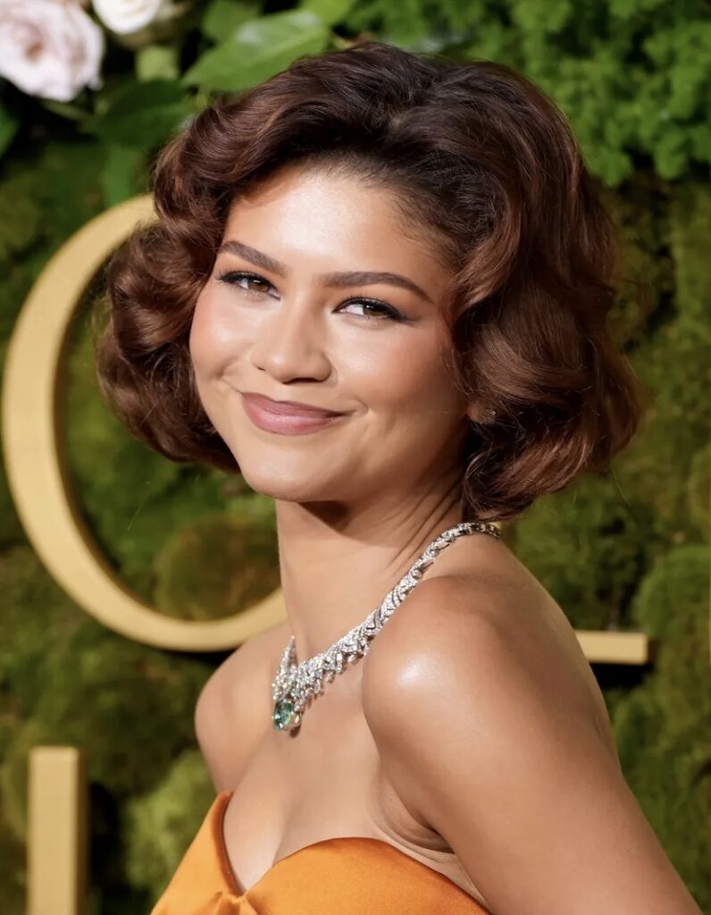 Zendaya gülümseyen dalgalı kahverengi saçlar elmas zümrüt kolye turuncu elbise