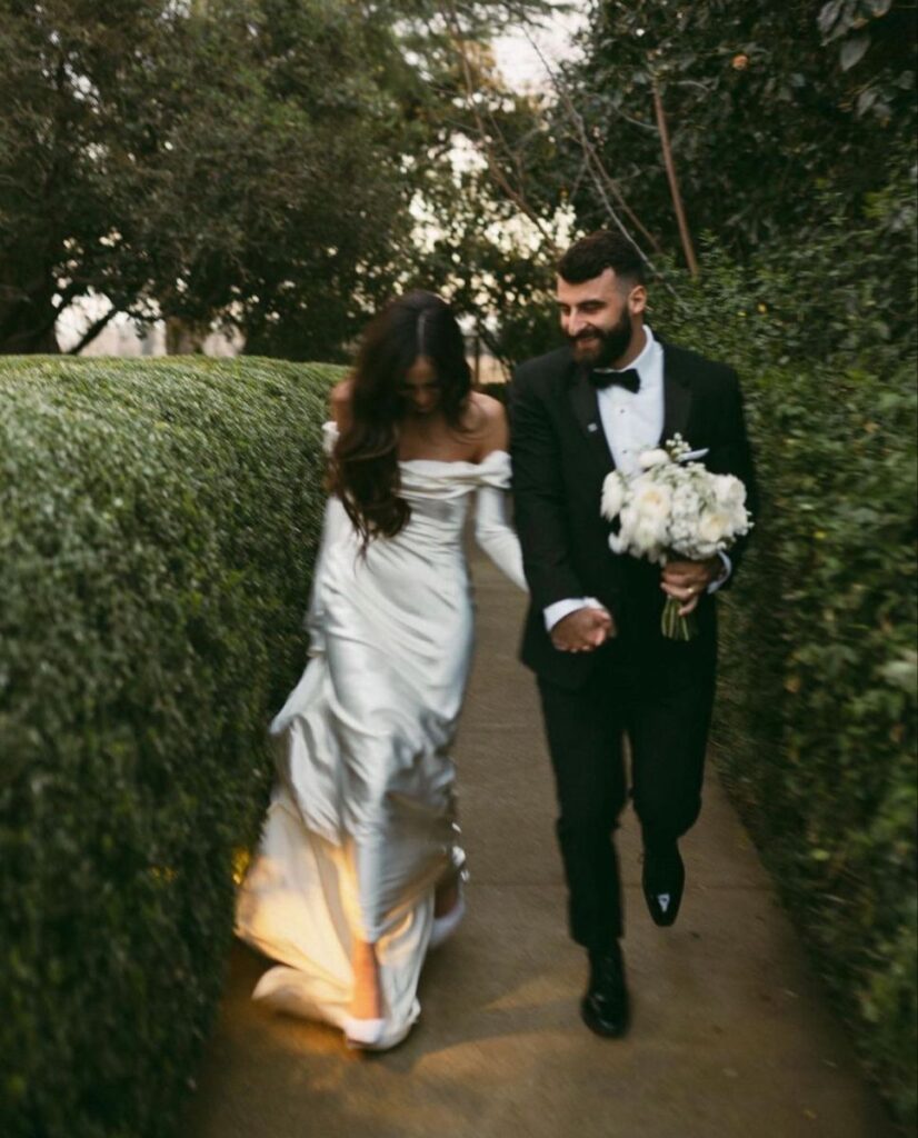 Bahçede el ele yürüyen gelin ve damat, damat beyaz buket tutuyor